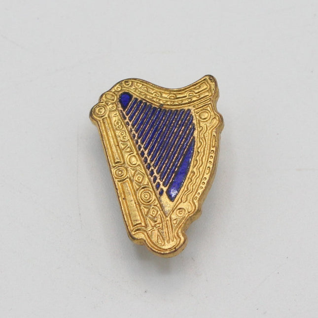 A small gold-toned harp-shaped Guinness pin badge with a dark blue inlay, photographed on a plain light background.