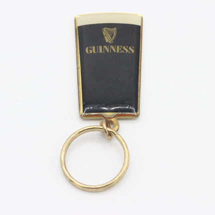 A vintage Guinness pint glass keychain with a black enamel body, gold-plated metal border, gold lettering, and a gold split key ring, shown on a white background.