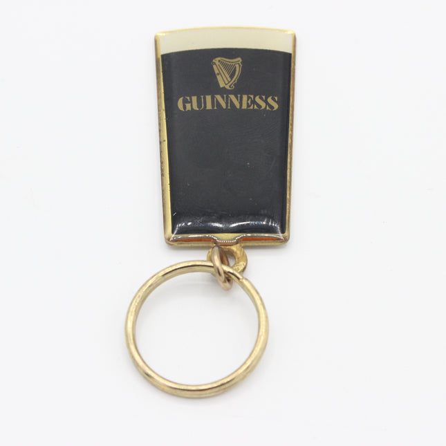A vintage Guinness pint glass keychain with a black enamel body, gold-plated metal border, gold lettering, and a gold split key ring, shown on a white background.