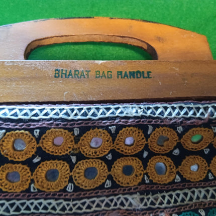Close-up of a wooden bag handle with the text 'BHARAT BAG HANDLE' printed on it, resting on a bright green textured surface. The handle is attached to a black fabric body decorated with rows of gold circular mirror work and white geometric embroidery.