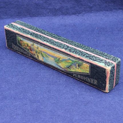 A vintage M. Hohner Rheingold” Harmonica featuring a textured black and green patterned top, a colorful landscape illustration, and gold lettering, resting on a blue textured background.