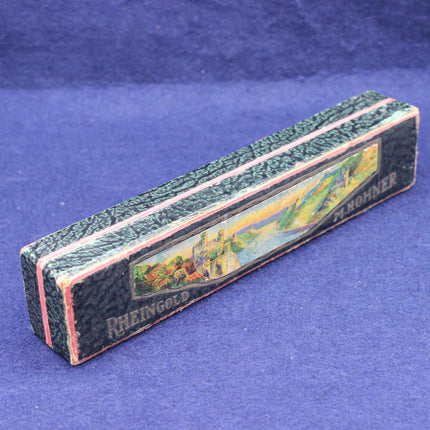 A vintage rectangular box with a textured dark green and black patterned exterior, pink trim, and a colorful landscape illustration on the front. The words 'RHEINGOLD' and 'RHOHNER' are printed on the box, which is photographed on a textured blue background.