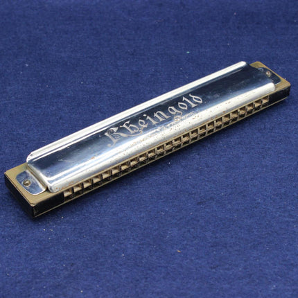 A vintage-style harmonica with a polished silver metal body and gold-toned ends, featuring the brand name 'Rheingold' engraved on the top, resting on a textured blue fabric background.