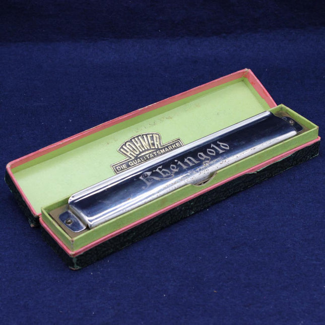 A silver metal harmonica in an open vintage presentation box with a green interior and pink trim, placed on a dark blue textured background. The brand name 'Hohner' is visible on the inside lid.