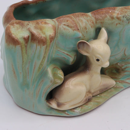A ceramic planter shaped like a tree trunk with a cream-colored fawn figurine sitting in the foreground, featuring a glossy turquoise and brown glaze.