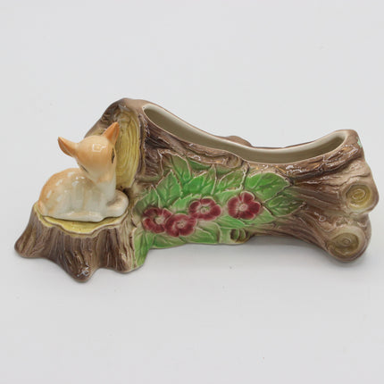 A decorative ceramic planter shaped like a tree stump with a small cream-colored deer figurine sitting on the left side. The planter features painted green leaves and red flowers along the side, set against a plain light gray background.