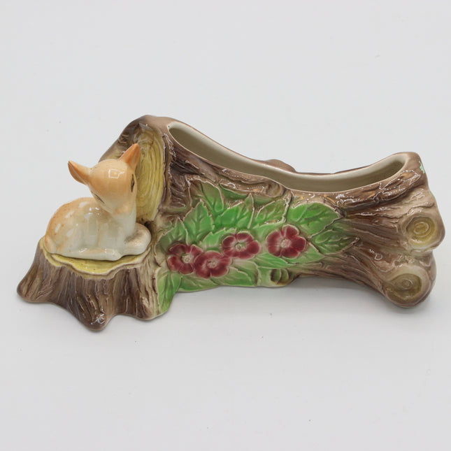 A decorative ceramic planter shaped like a tree stump with a small cream-colored deer figurine sitting on the left side. The planter features painted green leaves and red flowers along the side, set against a plain light gray background.