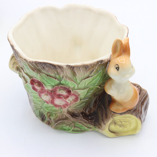 A ceramic cup shaped like a tree trunk with a small rabbit figurine perched on the side, featuring green leaves, pink flowers, and a cream-colored interior, shown on a white background.