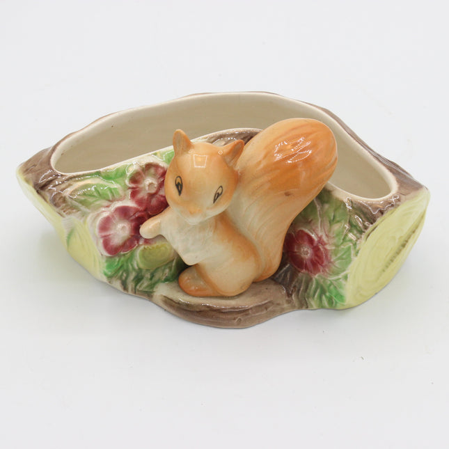 A ceramic squirrel planter shaped like a tree trunk, featuring a glossy orange-brown squirrel figurine sitting in the center, with painted green leaves and pink flowers around the base, set against a plain white background.