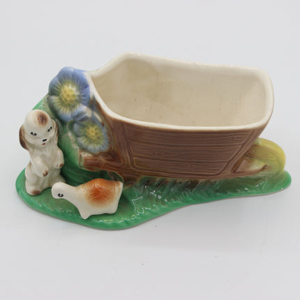 A vintage ceramic basket planter shaped like a brown basket on a green grassy base with two small animal figures and blue flowers, photographed on a white background.