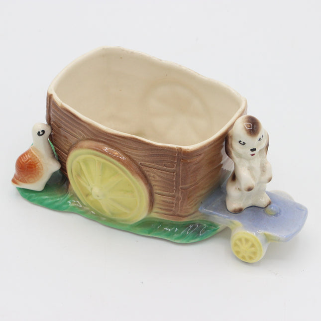 A ceramic planter shaped like a small brown cart with yellow wheels, green grass, a blue platform, and two small animal figurines on a plain white background.