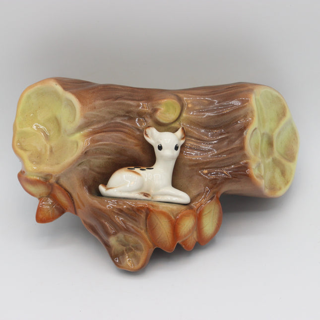 A ceramic figurine of a white deer nestled inside a brown and tan log-shaped base with pale green inner ends, photographed on a light gray background.