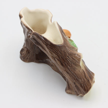 A ceramic tree stump planter with a textured brown bark exterior and a glossy cream-colored interior, featuring a small green leaf and orange acorn detail on the side, set against a plain white background.
