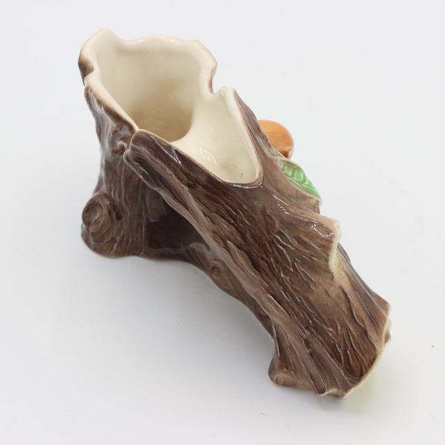 A ceramic tree stump planter with a textured brown bark exterior and a glossy cream-colored interior, featuring a small green leaf and orange acorn detail on the side, set against a plain white background.