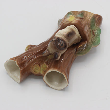 A ceramic decorative figurine shaped like a tree stump with two hollow openings at the ends and a small animal figure nestled inside, shown on a plain light background.
