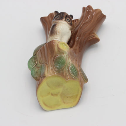 A ceramic figurine of a squirrel perched on a tree stump with green leaves, shown on a plain white background.