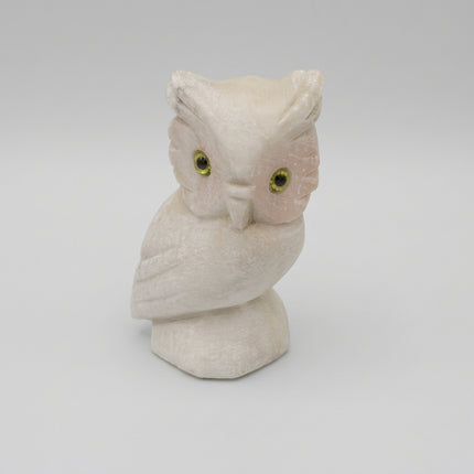 A small carved owl figurine in a warm ivory or cream stone-like material, with yellow eyes, standing on a matching base against a plain light gray background.