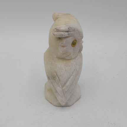 A small carved stone owl figurine in a light cream or off-white color, featuring a small green stone eye, set against a plain light gray background.