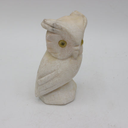 A small carved owl figurine made of a textured off-white material, featuring yellow-green eyes, set against a plain light gray background.