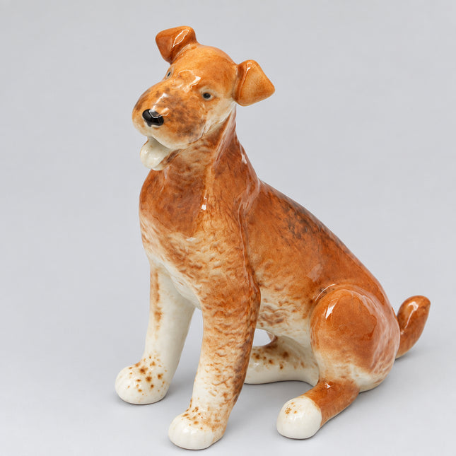 A glossy ceramic figurine of a sitting dog in shades of brown, tan, and cream, photographed against a seamless white background.