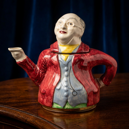 A ceramic teapot shaped like a man in a red coat pointing outward, displayed on a polished wooden surface against a dark blue curtain background.