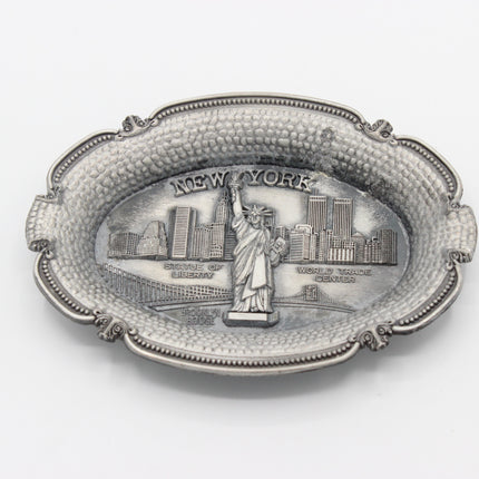 An oval silver-toned metal tray featuring an embossed relief of the Statue of Liberty and the New York City skyline, with the words 'NEW YORK' across the top, shown on a plain white background.