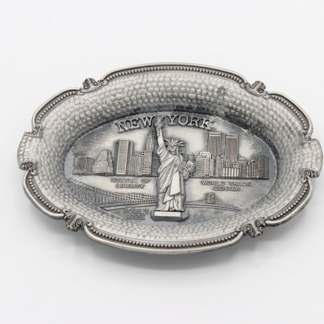 An oval silver-toned metal tray featuring an embossed relief of the Statue of Liberty and the New York City skyline, with the words 'NEW YORK' across the top, shown on a plain white background.