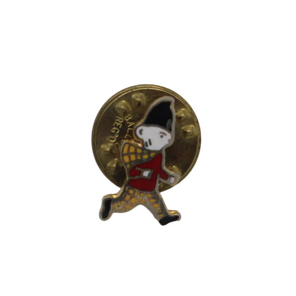 A small enamel pin featuring a walking character in a red coat and yellow patterned pants, set against a gold circular background, on a plain light gray surface.