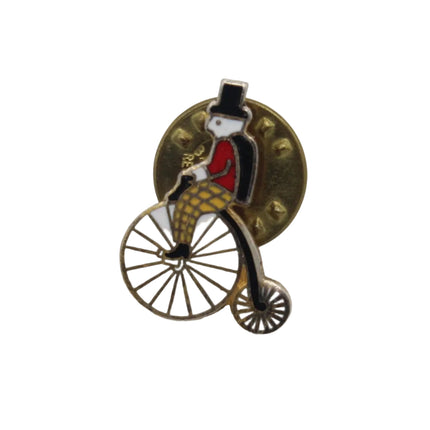 A vintage-style penny-farthing bicycle enamel pin with a gold-toned metal finish, featuring Rupert Bear in a red jacket and black top hat on a plain white background.