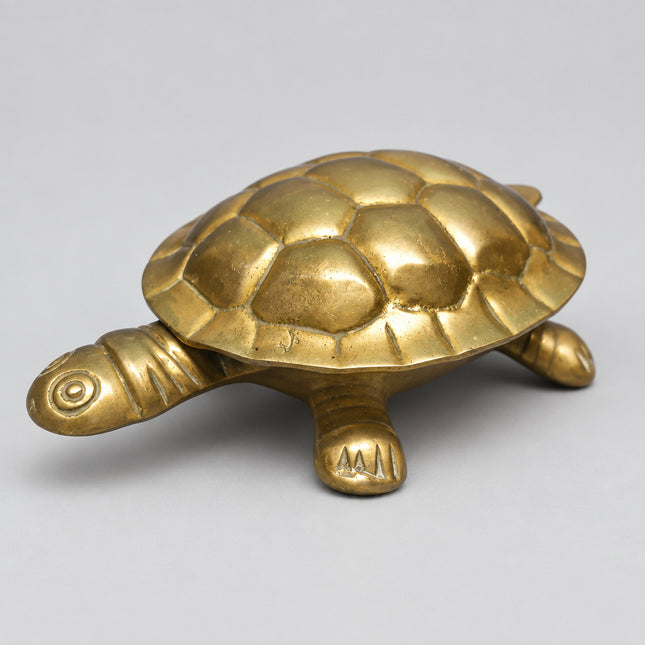 A small metallic turtle figurine with a polished brass or gold finish, photographed against a solid green background.