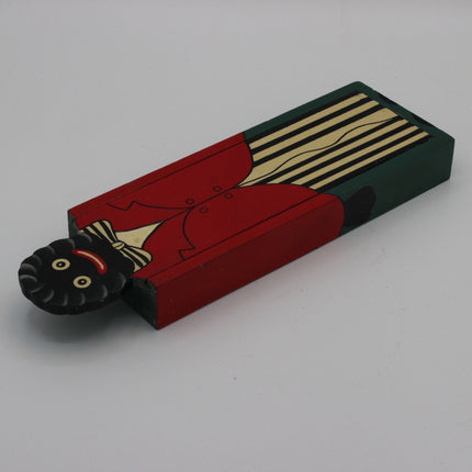 A hand-painted wooden toy or decorative block featuring a stylized figure with a black face, red coat, cream details, and a dark green striped background, photographed on a plain light gray surface.