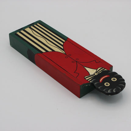 A hand-painted wooden pencil box with a black and white face peeking out from the side. The matchbox features a red coat, cream stripes, and a dark green background.