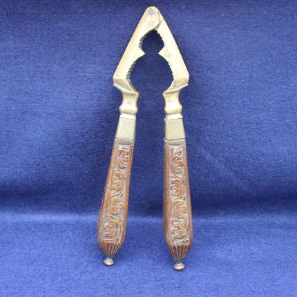A vintage nutcracker with ornate carved brown wooden handles and a gold-toned metal head, displayed on a textured blue fabric background.