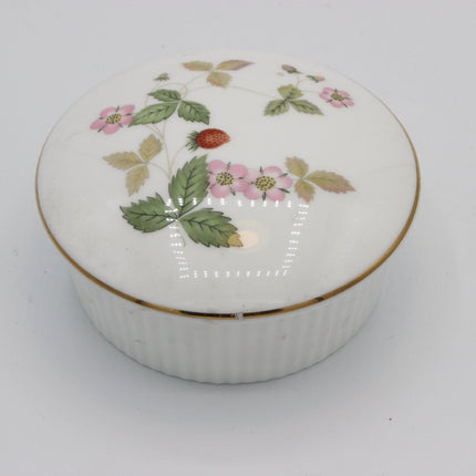 A round white porcelain lidded box with a delicate hand-painted strawberry and leaf pattern on the lid, accented by a thin gold band at the rim, set against a plain light gray background.