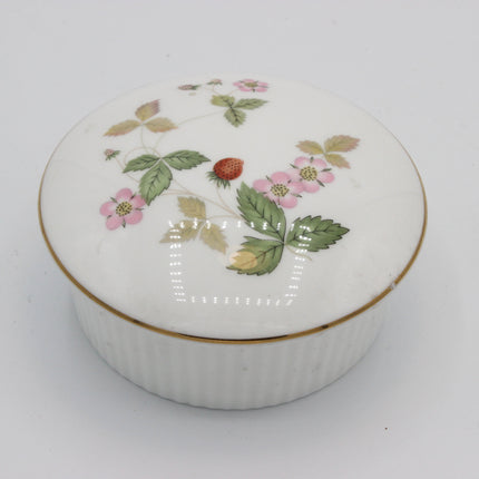A round white porcelain keepsake box with a matching lid, decorated with a hand-painted strawberry and pink flower motif and a thin gold trim, set against a plain light gray background.