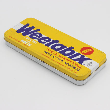 A vintage yellow Weetabix Pencil Box tin for Woolworths, featuring the brand name in blue and red text on a yellow background, photographed on a light gray surface.