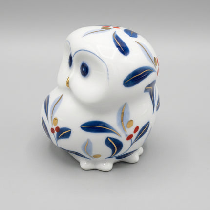 A small ceramic owl figurine or container in glossy white, decorated with blue leaf motifs and small gold and red accents, photographed on a plain white background.