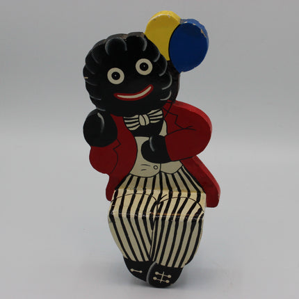 A hand-painted wooden Golly figure featuring a black body, red jacket, cream-and-black striped pants, and balloons in yellow and blue, set against a plain gray background.