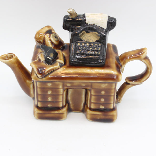 A ceramic novelty teapot shaped like a typist at a desk, featuring a brown and amber glazed body with a black typewriter figure on top, photographed against a plain white background.