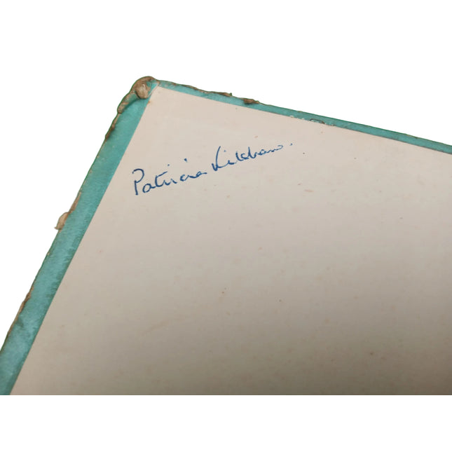 A close-up of a vintage book or notebook corner with a cream-colored page, a light blue cloth binding, and a blue handwritten signature reading 'Patricia Kildian' on a green felt-like surface.