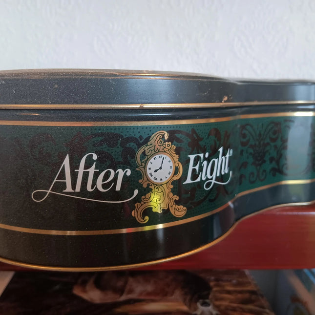 A vintage After Eight tin canister with a dark green and black body, gold trim, and a central gold clock illustration, resting on a wooden shelf against a white wall.
