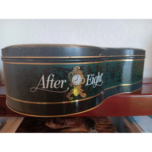 A vintage After Eight tin canister with a dark green and black body, gold trim, and a central gold clock illustration, resting on a wooden shelf against a white wall.