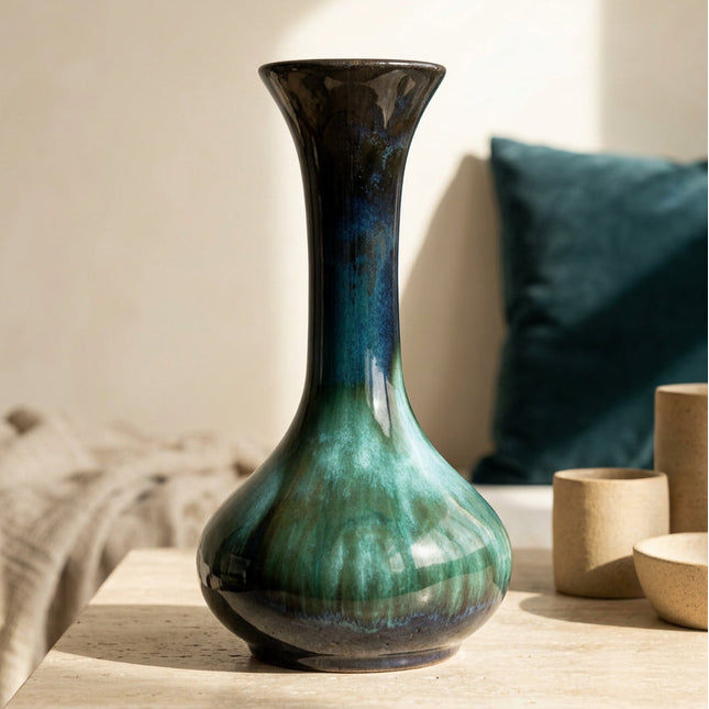 A ceramic bud vase with a flared rim and a bulbous base, featuring a glossy glaze that transitions from deep black at the top to rich teal and emerald green at the bottom, in a modern living room setting.