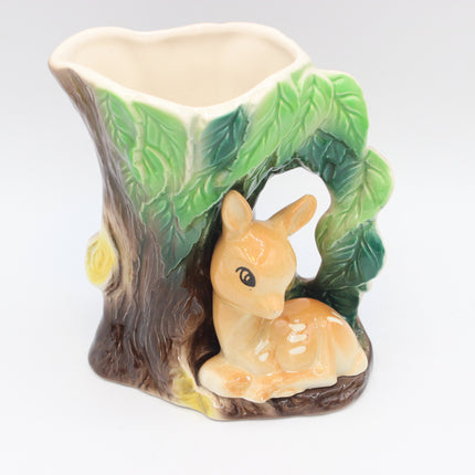 A ceramic woodland-themed cup or planter shaped like a tree stump with green leaves, a brown trunk, and a small cream-colored deer figurine sitting in front, shown on a plain light background.