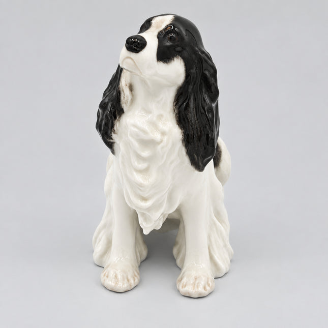 A ceramic figurine of a black and white long-eared dog, likely a Cocker Spaniel, sitting down, photographed against a plain white background.