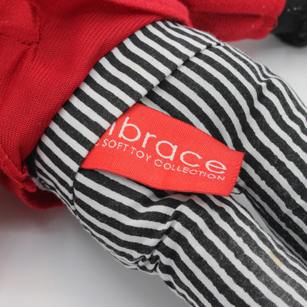 Close-up of a soft Golly toy wearing black and white striped pants and a red top, with a visible red fabric tag reading "embrace SOFTTOY COLLECTION.".