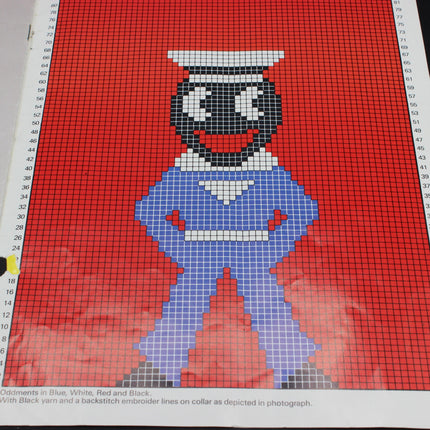 Cross-stitch pattern of a Golly Sailor character in blue and white on red fabric with a ruler for scale.