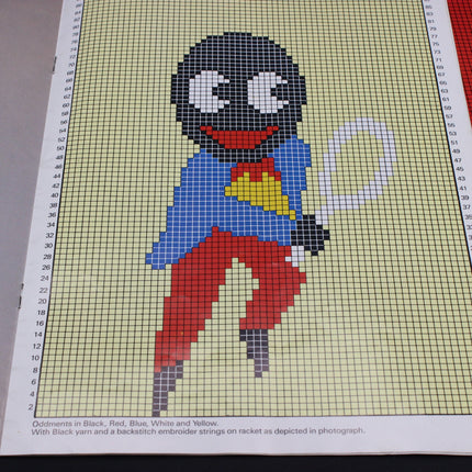 Cross-stitch pattern of a Golly Tennis character with a black head, blue shirt, red pants, and yellow heart on a grid background.