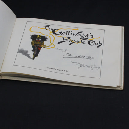 Book open to a page with a illustration of a character on a bicycle and text 'The Gollivogg's Bicycle Club' on a black background.