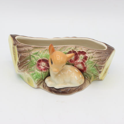 A ceramic planter shaped like a tree trunk with a small white and tan deer figurine in the center, flanked by red flowers and green leaves, on a plain white background.
